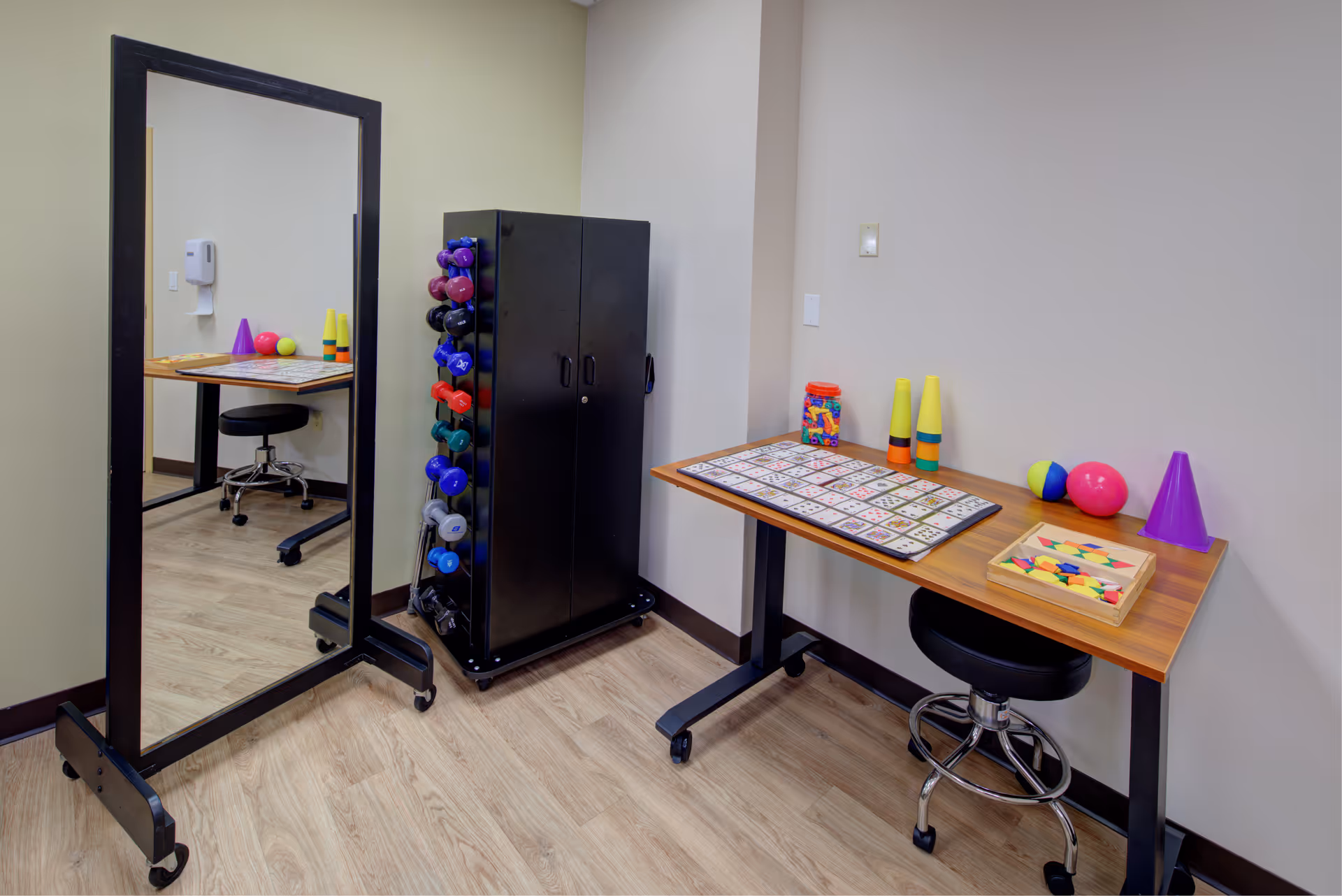 A therapy or activity room with a large standing mirror, a black cabinet holding colorful dumbbells, and a wooden table with various therapy and sensory items including a bingo card, stacking cones, balls, and a box of colorful shapes. A black rolling stool is positioned under the table.