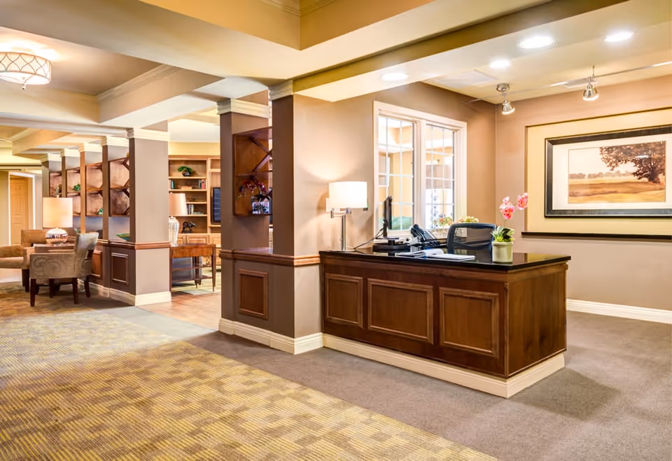 Warmly lit reception desk and adjacent seating/lounge area in a senior living facility lobby with built-in shelves and artwork.