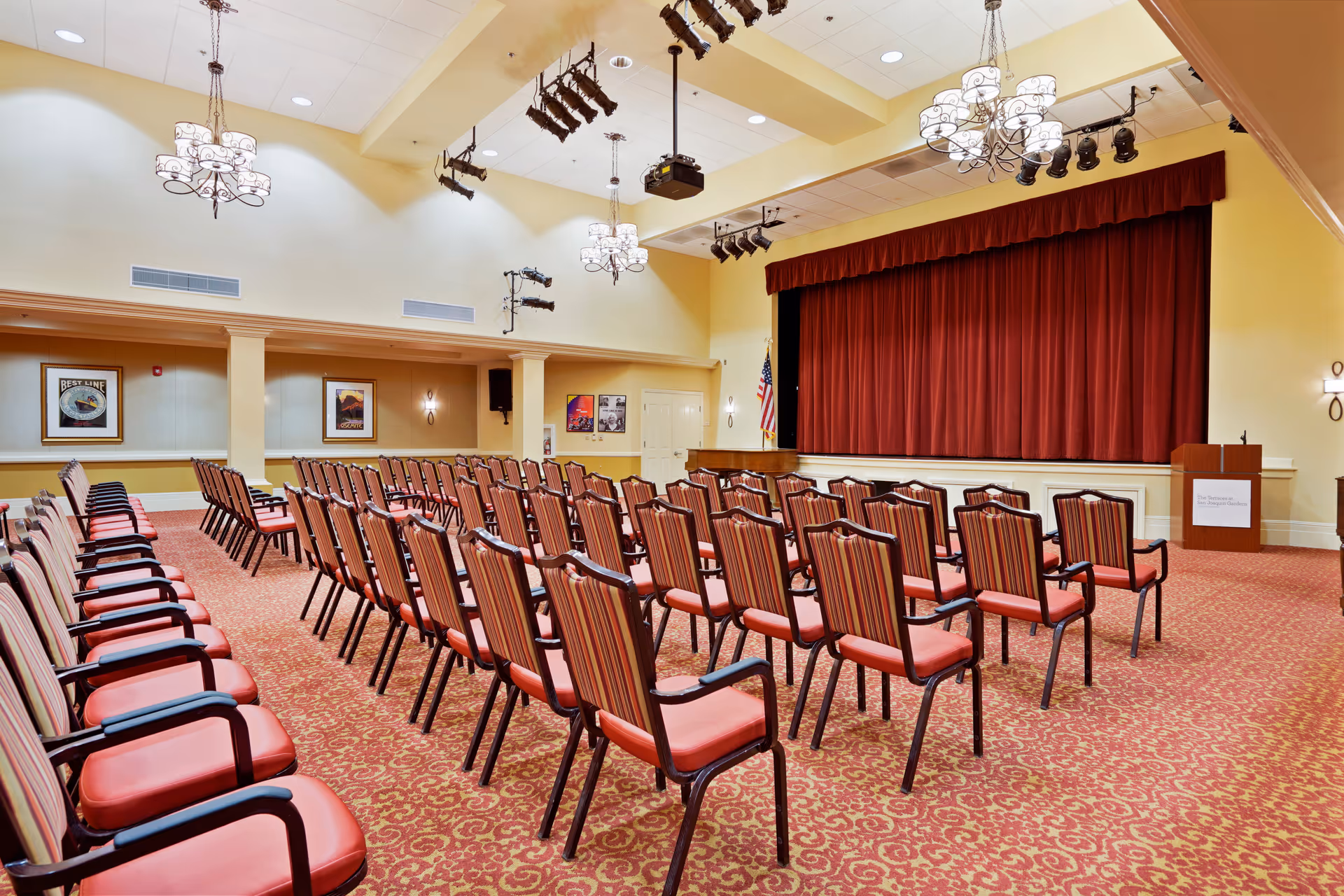 A large auditorium with rows of red cushioned chairs facing a stage with a closed red curtain. The room has yellow walls, ornate chandeliers, stage lighting, and framed pictures on the walls. There is a podium on the right side of the stage with a sign that reads 'The Terraces at San Joaquin Gardens'.