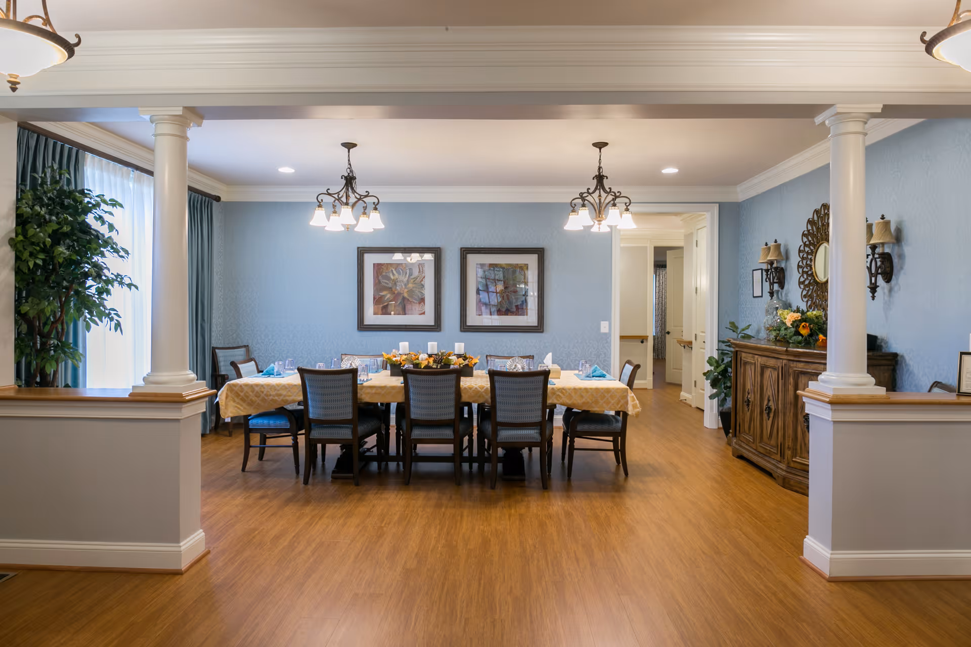 A well-lit dining room with a long table covered with a yellow tablecloth, set with plates, glasses, and napkins. The room features wooden flooring, two chandeliers hanging from the ceiling, two framed artworks on a light blue wall, and a wooden sideboard with decorative items and a mirror. There are white columns framing the entrance to the dining area and a large plant near a window with blue curtains.
