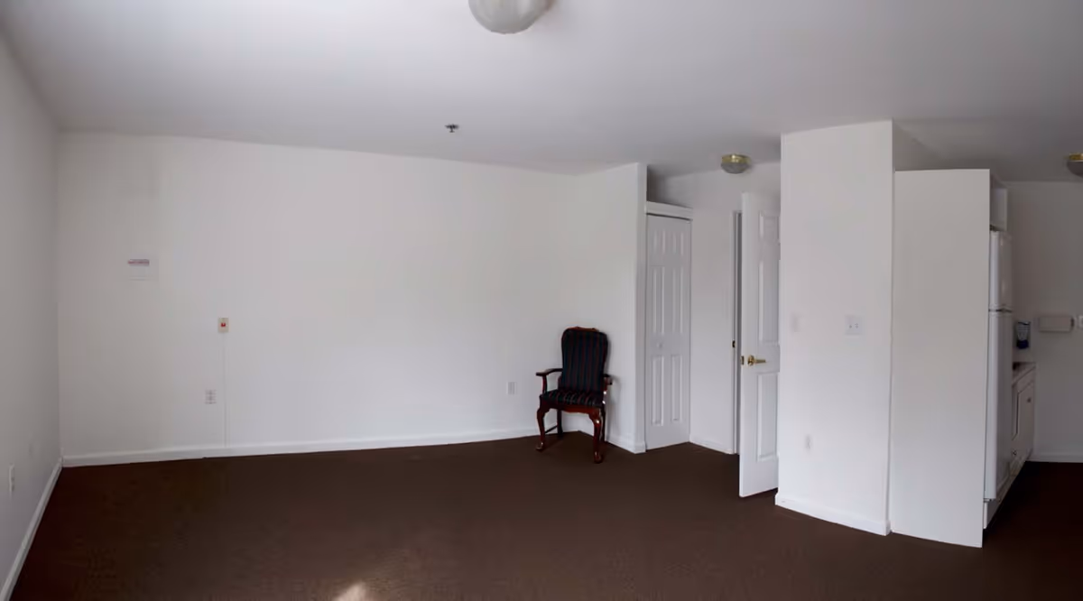 An empty room with white walls and brown carpet flooring. There is a single dark wooden chair with a cushioned seat and backrest placed near a corner. The room has two white doors, one slightly open, and a small kitchenette area with a refrigerator and cabinets on the right side.
