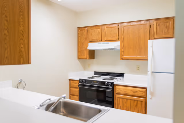 Small kitchen with oak cabinets, white countertops, a stainless steel sink in the foreground, an electric stove and a white refrigerator.