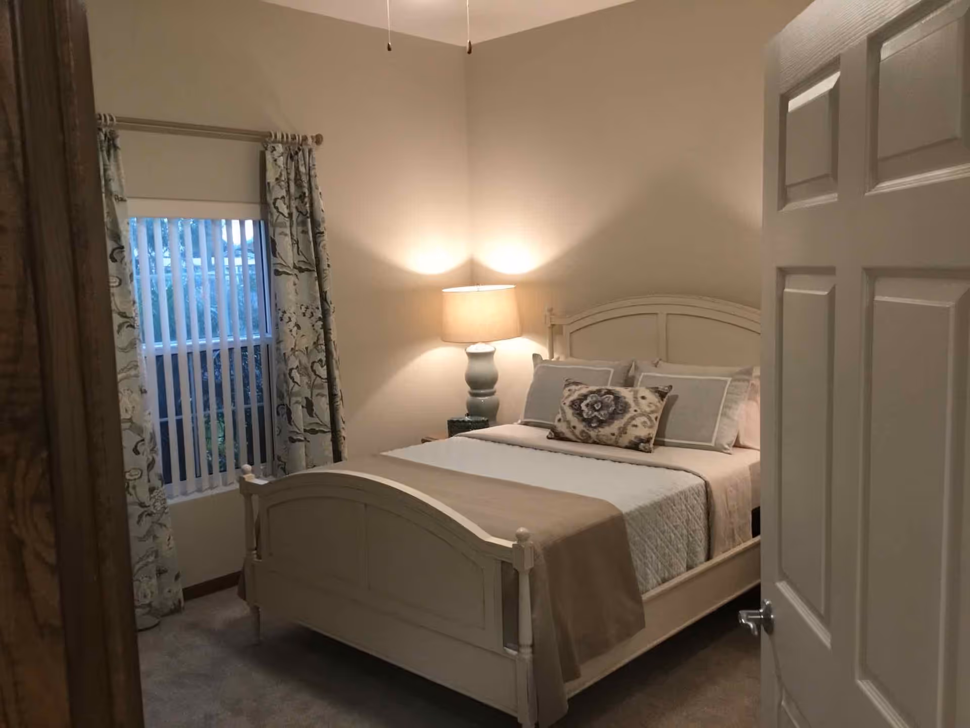 A cozy bedroom with a white wooden bed frame, neatly made with beige and white bedding and decorative pillows. A bedside table with a lamp is lit, casting a warm glow on the beige walls. There is a window with vertical blinds and floral curtains on the left side of the room. The door to the room is partially open.
