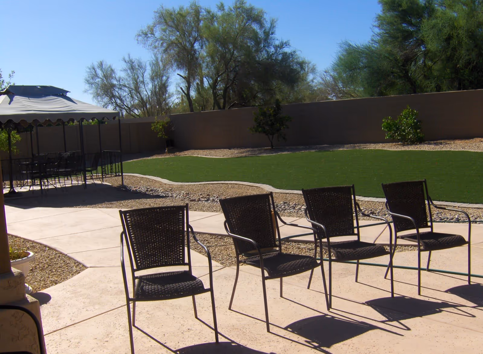 Sunlit outdoor courtyard with a row of metal patio chairs, a shaded gazebo, and a lawn area bordered by a block wall and trees.