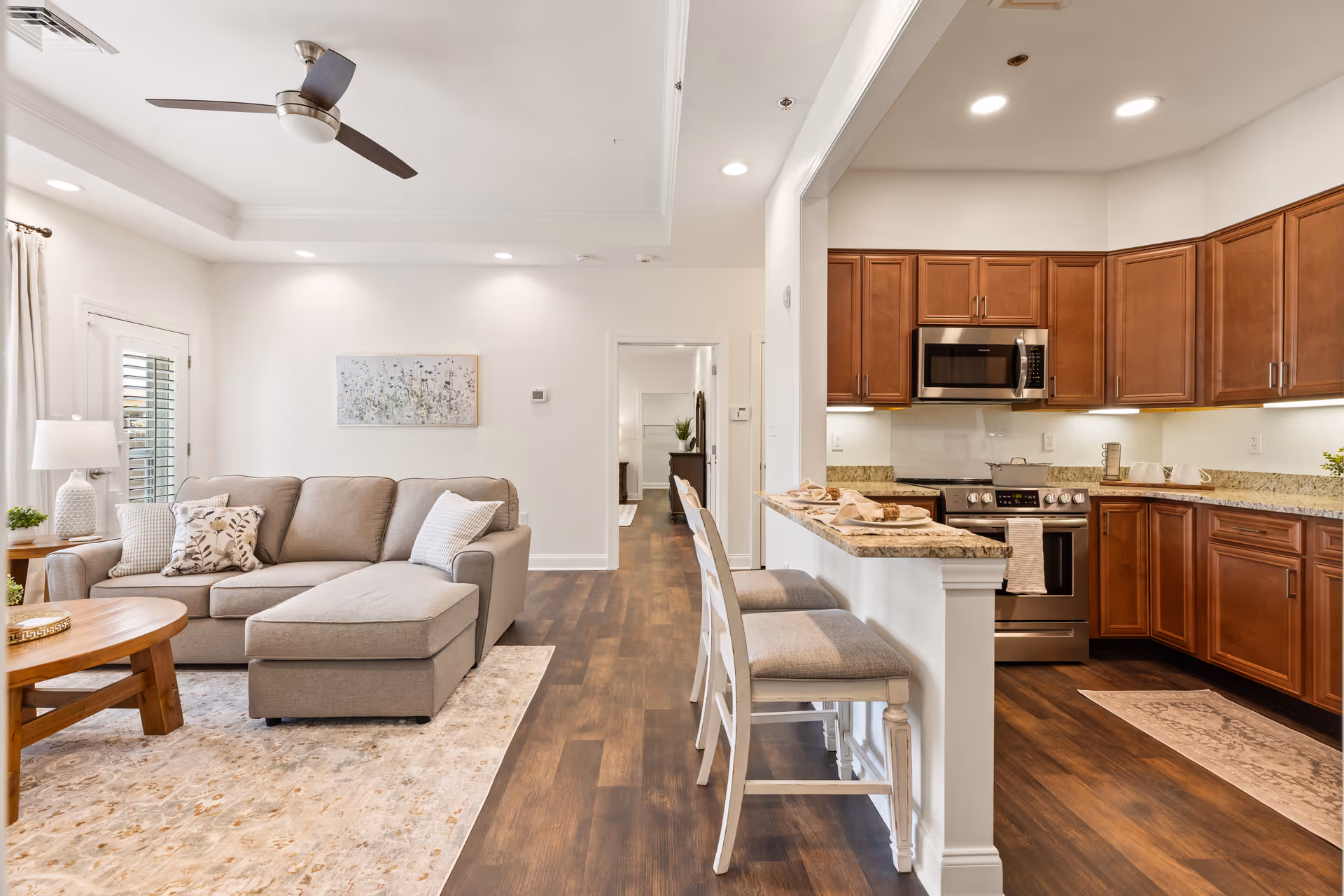 A bright and spacious living room and kitchen area in a senior living facility. The living room features a beige sectional sofa with patterned cushions, a wooden coffee table, a side table with a lamp and plant, and a ceiling fan. The kitchen has wooden cabinets, granite countertops, stainless steel appliances including a microwave and stove, and a breakfast bar with two cushioned stools. The flooring is a dark wood laminate, and the walls are painted white with recessed lighting.