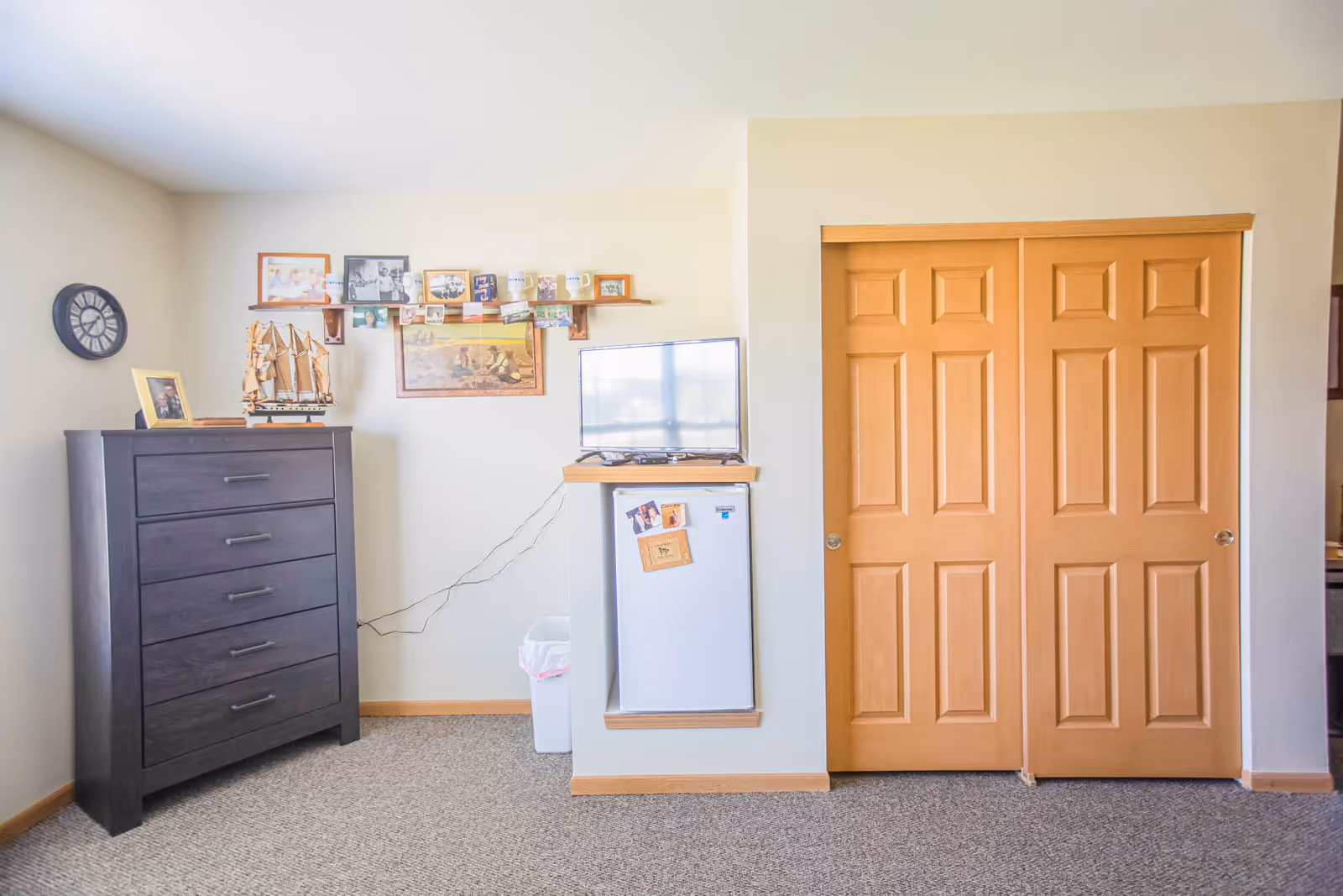 A room with beige walls and carpeted floor featuring a dark wooden dresser on the left, a small white refrigerator with a TV on top in the center, and wooden double sliding closet doors on the right. Above the refrigerator is a shelf with framed photos, mugs, and a painting on the wall.