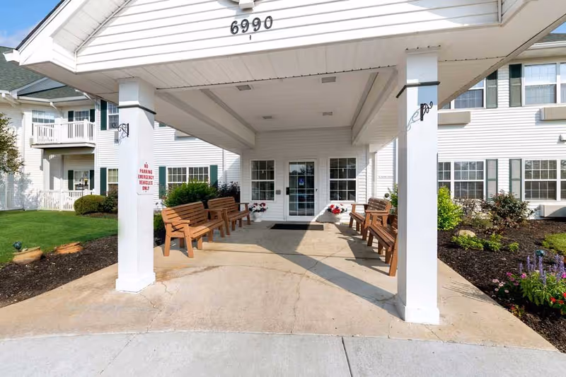 Entrance to a senior living facility with a covered drop-off area supported by white pillars. There are wooden benches on both sides under the canopy, flower pots near the glass double doors, and well-maintained landscaping with green grass and flower beds around the building. The building is white with multiple windows and a balcony visible on the left side. The address number 6990 is displayed above the entrance.