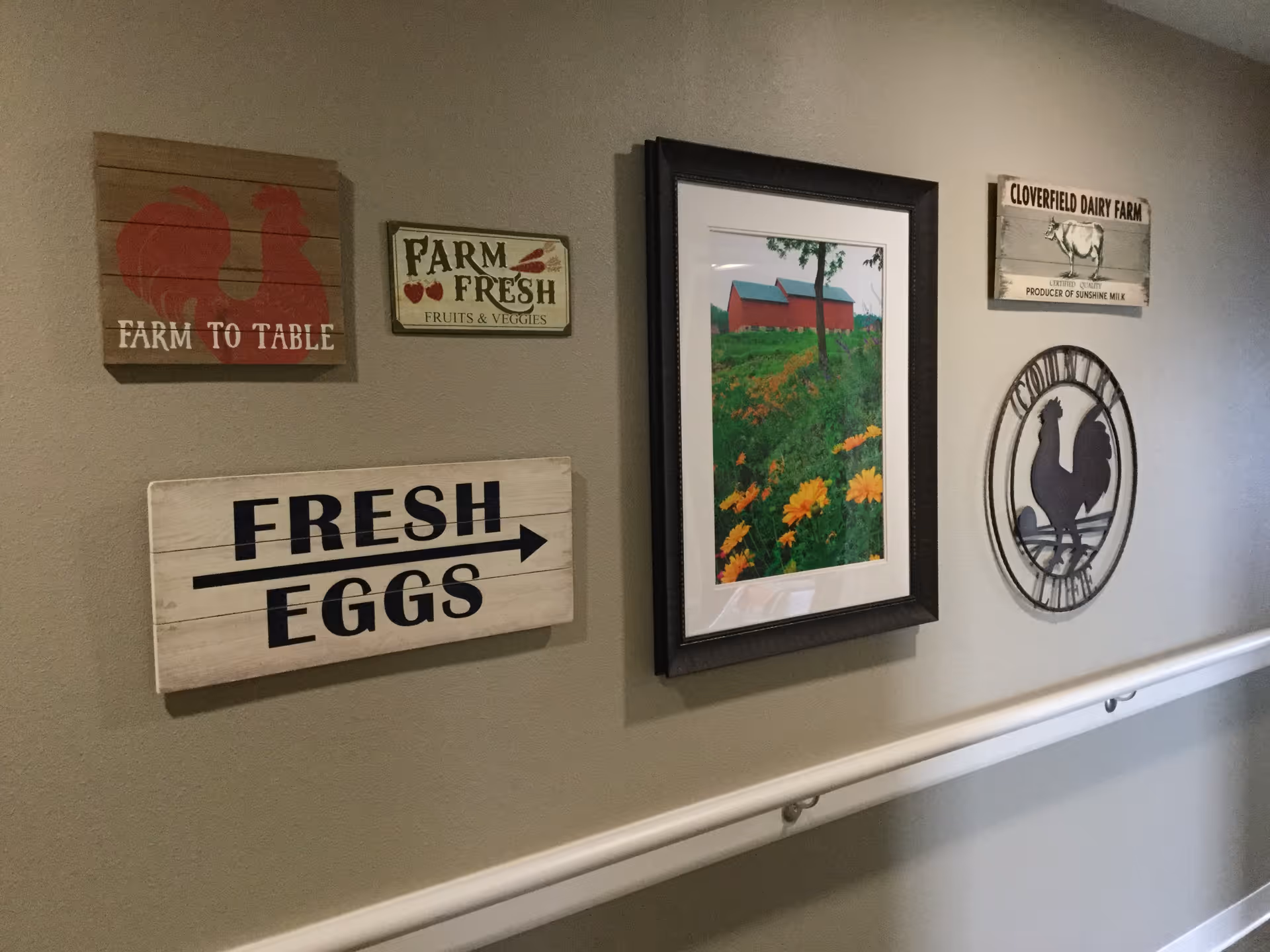 A hallway wall decorated with rustic farm-themed signs and artwork. The signs include a red rooster with the text 'FARM TO TABLE', a sign that says 'FARM FRESH FRUITS & VEGGIES', a directional sign reading 'FRESH EGGS', a framed picture of a red barn with a green field and yellow flowers, a sign for 'CLOVERFIELD DAIRY FARM' with a cow illustration, and a round metal rooster silhouette. A white handrail runs along the wall below the decorations.