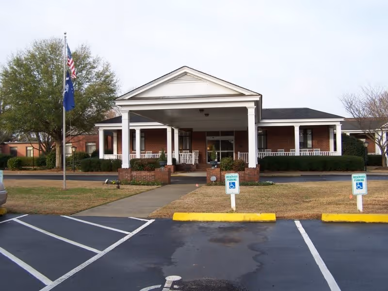 Front exterior view of The Florence Presbyterian Community building with a covered entrance supported by white columns, brick walls, a walkway leading to the entrance, two reserved handicapped parking spaces in front, and flags on a flagpole to the left.