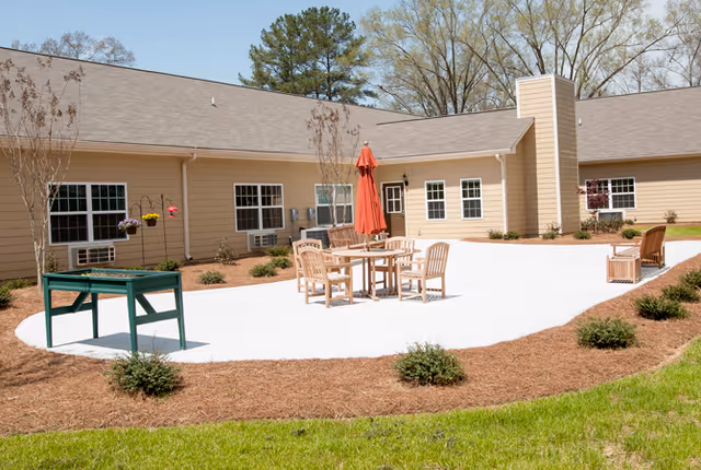 Outdoor patio area at a senior living facility with a round table, several wooden chairs, and a closed red umbrella. The patio is surrounded by mulch beds with small bushes and the beige exterior of the building with multiple windows is visible in the background.