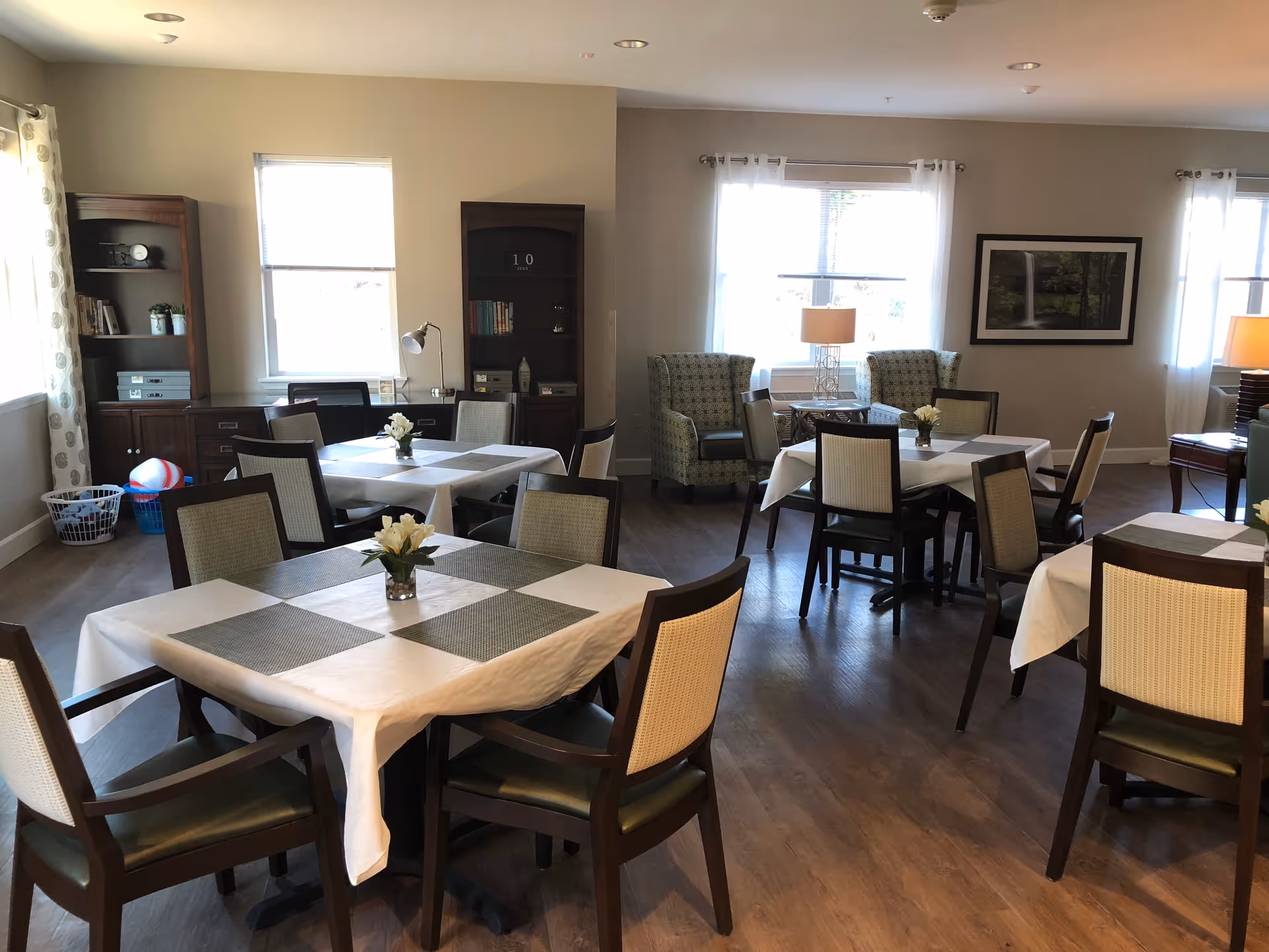 A well-lit dining room with several tables covered with white tablecloths and gray placemats, each table decorated with a small vase of white flowers. The room has wooden flooring, large windows with white curtains, two patterned armchairs near the windows, wooden bookshelves, and framed artwork on the wall.