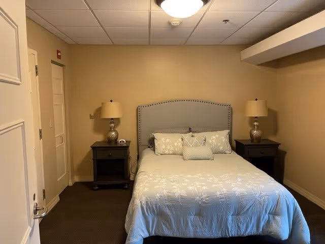 A bedroom with a neatly made bed featuring a light-colored bedspread and decorative pillows. The bed has a gray upholstered headboard with nailhead trim. On each side of the bed are dark wooden nightstands with matching table lamps. The walls are painted beige, and the ceiling has recessed lighting. There is a door on the left side of the room.