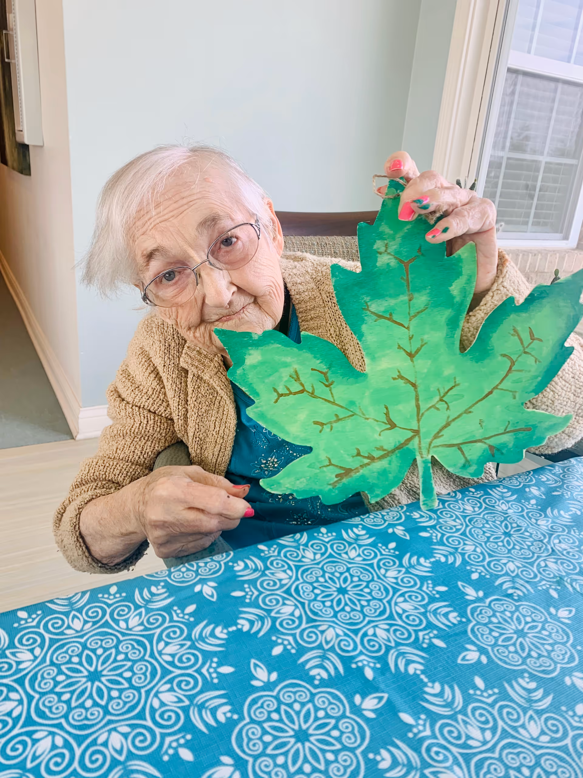 An elderly woman wearing glasses and a beige cardigan is sitting at a table covered with a blue patterned tablecloth. She is holding up a large green paper leaf with painted veins, showing it to the camera. Behind her is a light-colored wall and a window with white blinds.