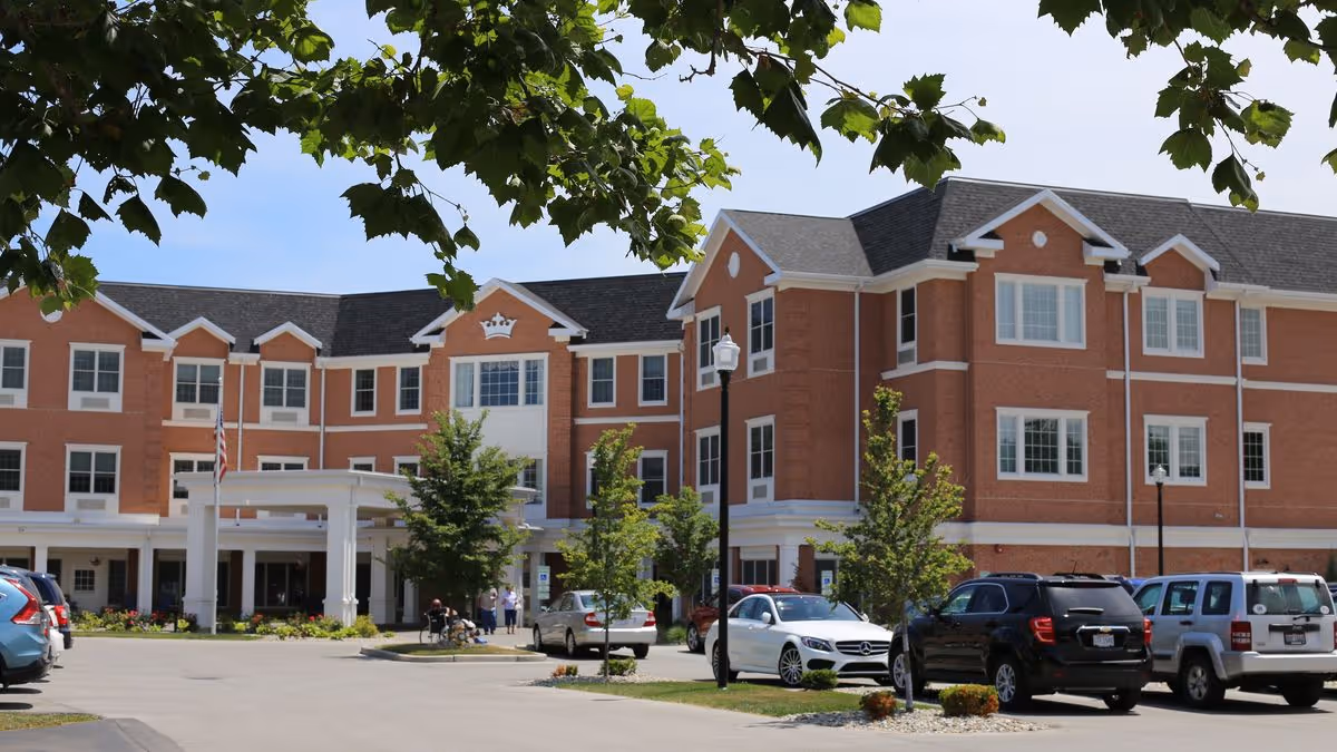 Exterior view of a three-story brick building with white trim, identified as Kingston Rehabilitation of Perrysburg. The building has multiple windows and a covered entrance with an American flag. Several cars are parked in front, and a few people are visible near the entrance. Trees and greenery surround the parking area under a clear sky.