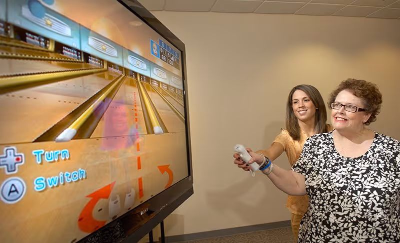 An elderly woman and a younger woman playing a virtual bowling game on a large screen using motion controllers in a room with beige walls.