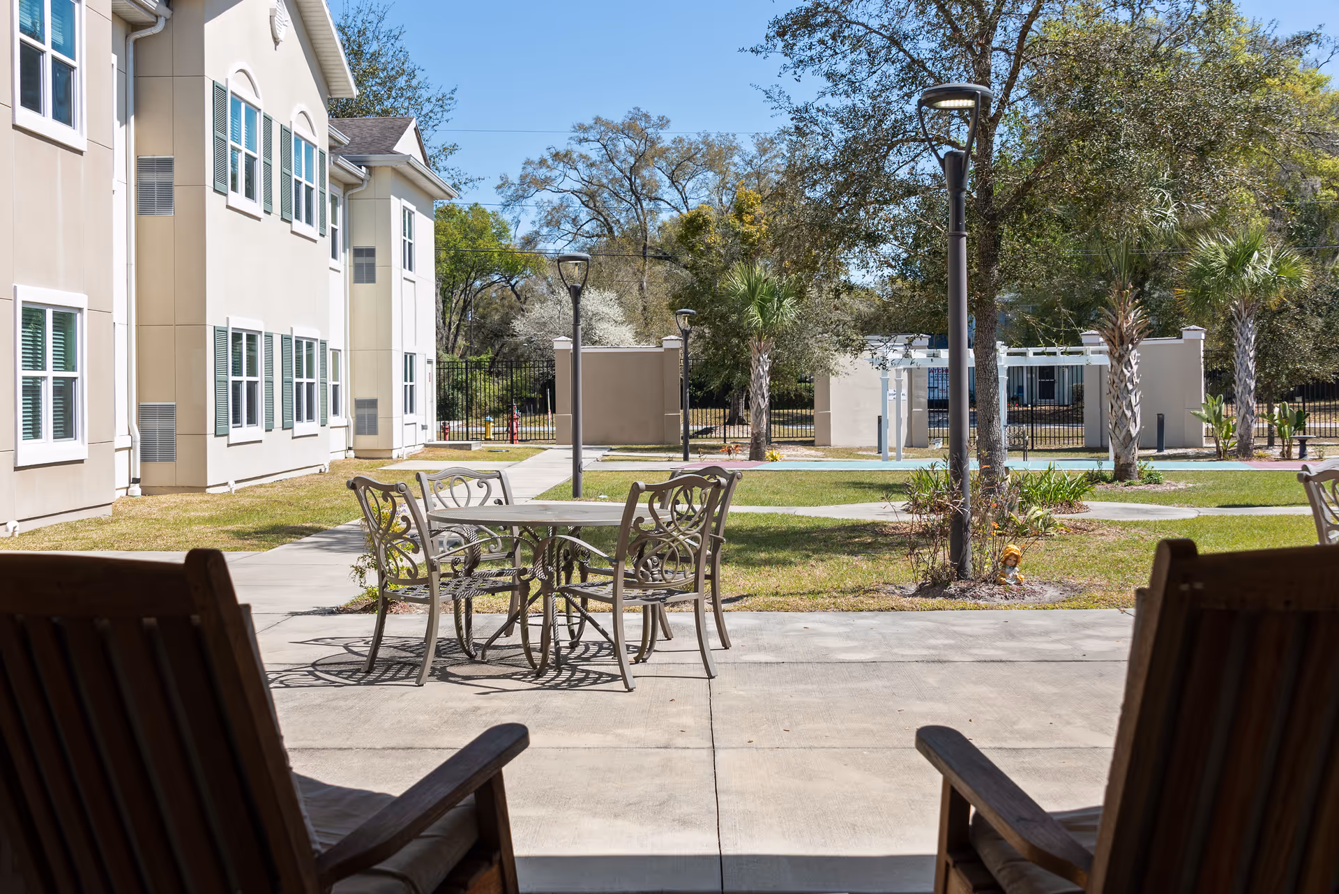 Outdoor patio area at Blue Palms Senior Living of Deland with metal table and chairs on a concrete surface, surrounded by grass, trees, and lampposts. The beige building with multiple windows is visible on the left side under a clear blue sky.