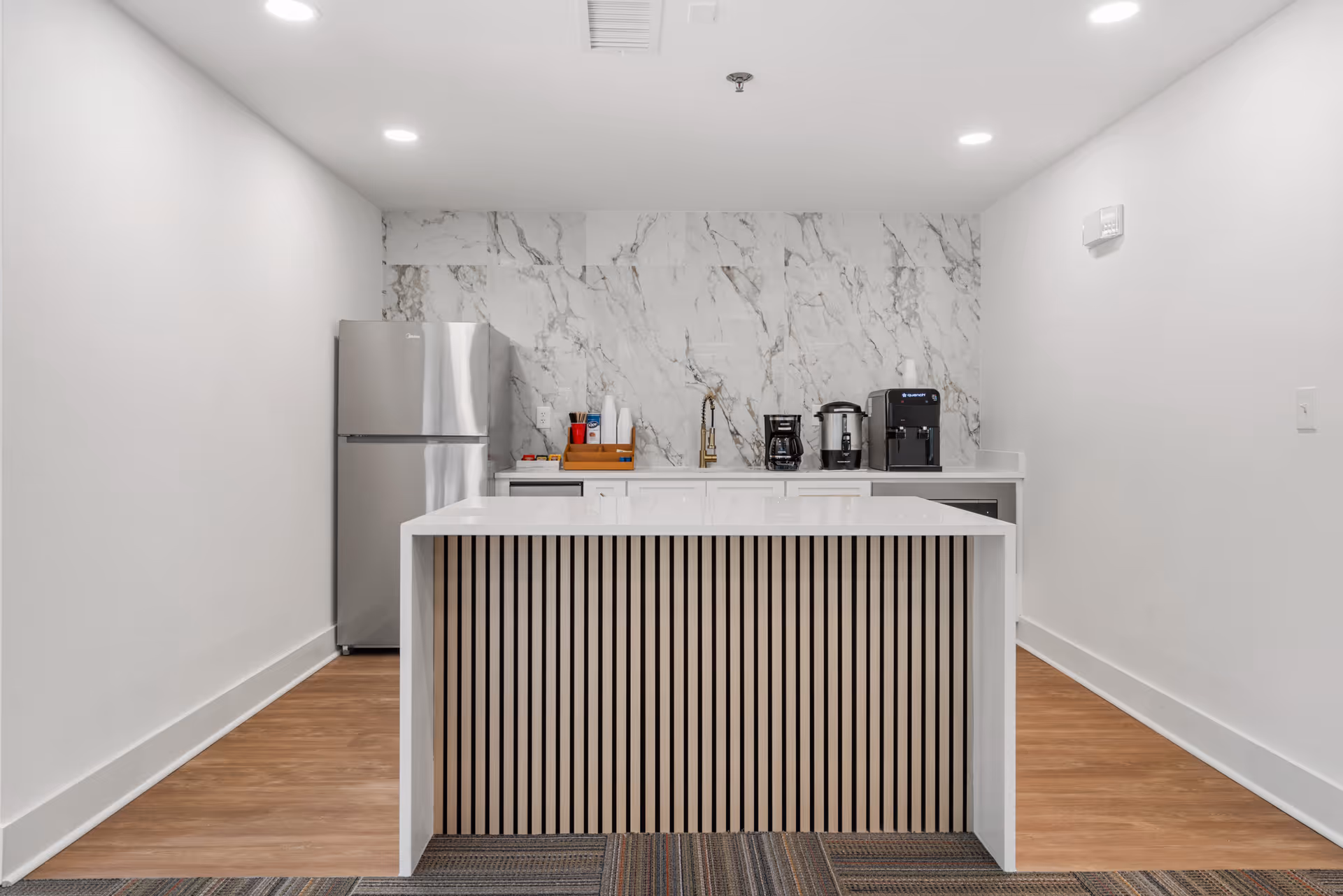 Small modern kitchenette with a white island, marble backsplash, stainless refrigerator and coffee machines.