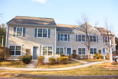 Exterior view of a two-story residential building with beige siding, multiple windows with white frames and blue shutters, and three blue front doors. There are small bushes and leafless trees in front of the building, along with a white picket fence section and a paved walkway leading to the doors. The sky is clear and blue.
