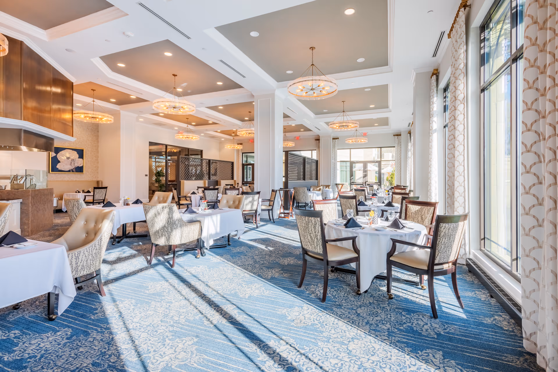 Bright, elegant dining room with tables dressed in white linens, upholstered chairs, large windows, and decorative chandeliers.