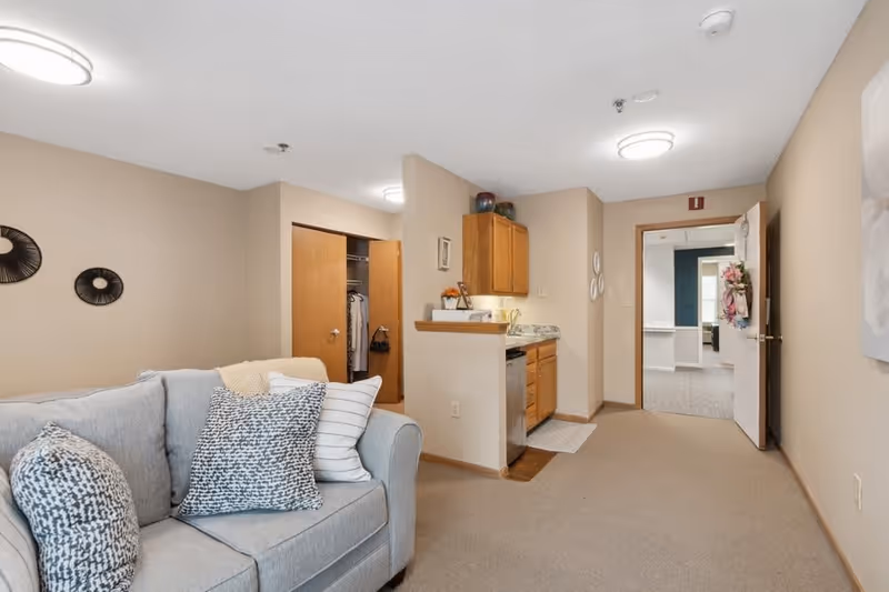 Interior view of a senior living facility apartment showing a light gray couch with patterned pillows on the left, a small kitchenette with wooden cabinets and a granite countertop in the center, and an open doorway leading to another room in the background. The walls are beige, and the floor is carpeted.