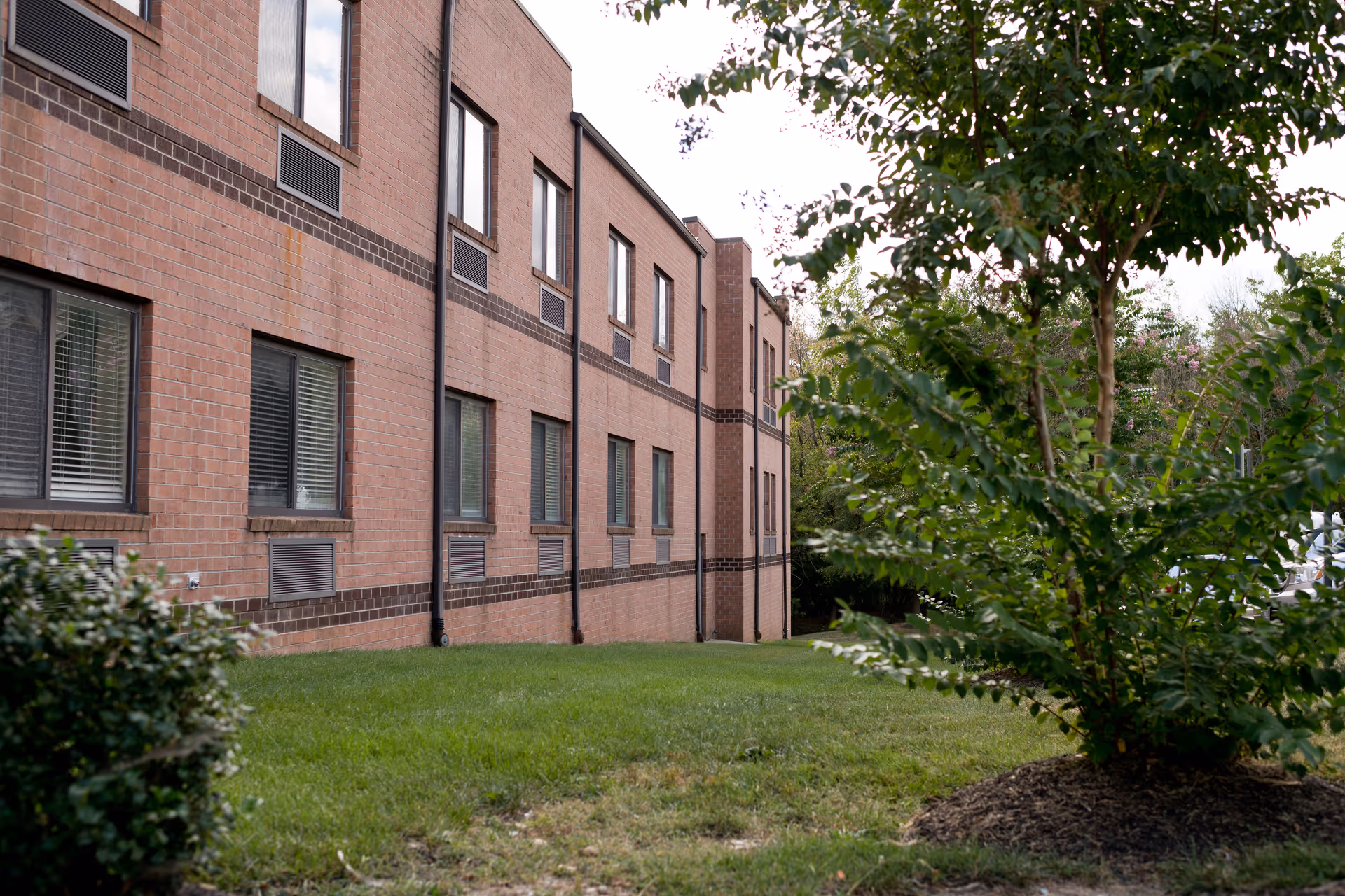 Exterior view of a two-story brick building with multiple windows, surrounded by green grass and trees.