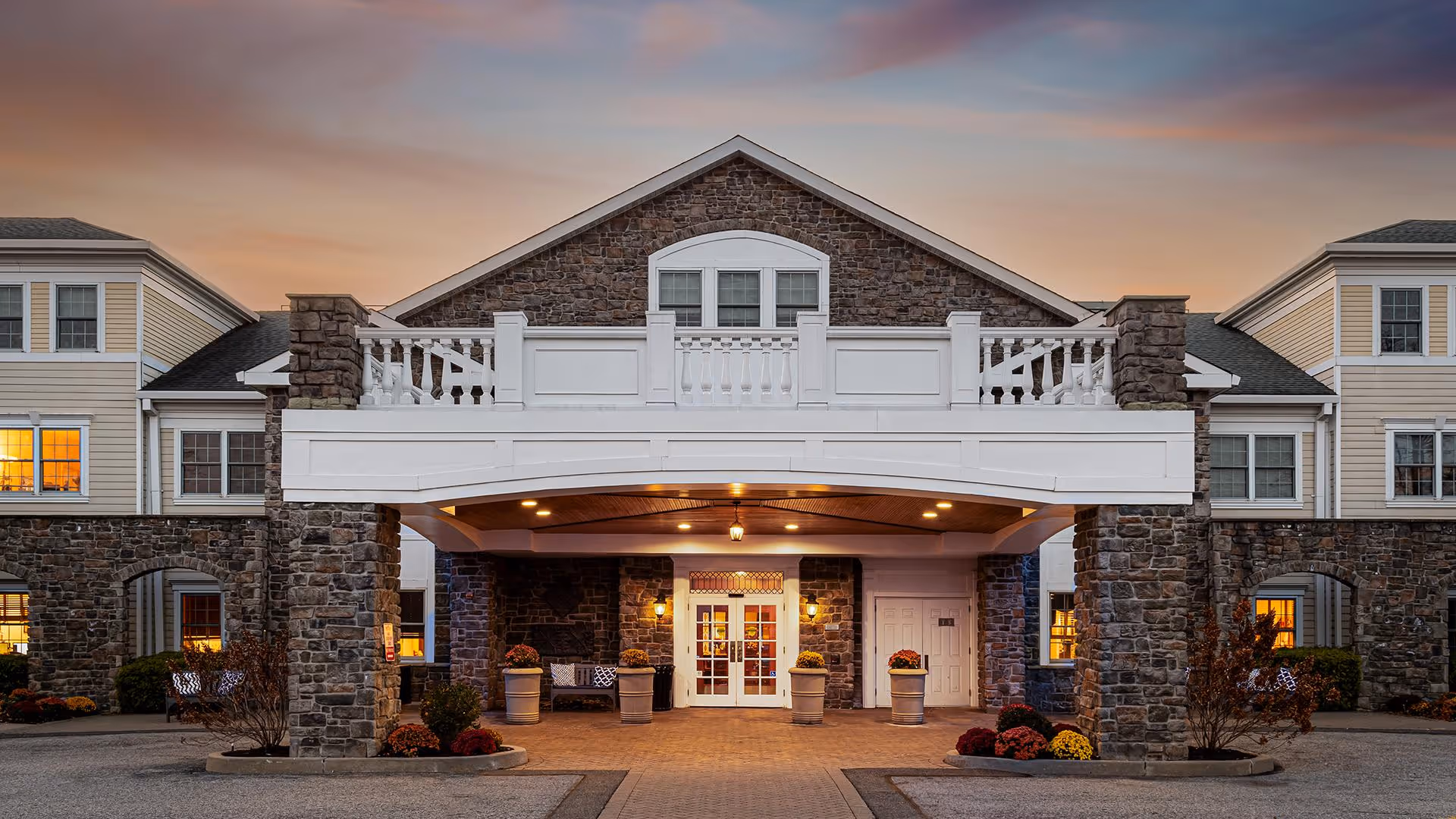 Front exterior view of Atria Rye Brook facility at dusk, featuring a stone and siding building with a covered entrance supported by stone pillars, illuminated by warm lights, and decorated with potted plants and benches.