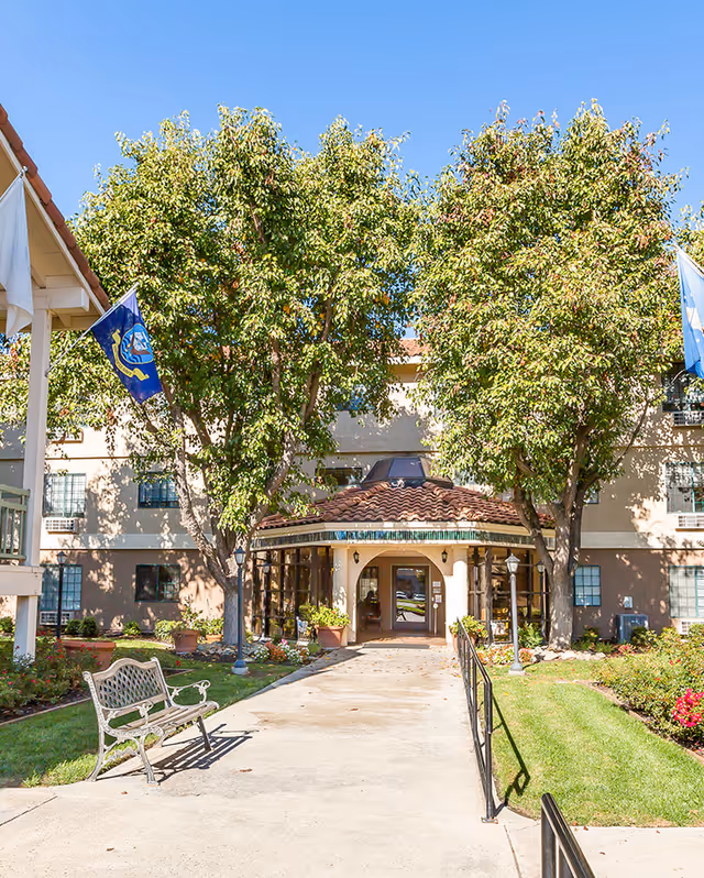 Entrance to a senior living facility with a tiled roof canopy, flanked by two large leafy trees. There is a concrete walkway leading to the door, a metal bench on the left, and well-maintained grass and flower beds on either side. Flags are visible on poles near the entrance.