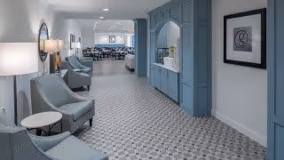 A bright and spacious hallway in a senior living facility with patterned tile flooring, light blue walls and built-in cabinetry. Several comfortable armchairs with small round tables are arranged along the left side. The hallway leads to a dining area with tables and chairs visible in the background. Wall sconces and framed artwork decorate the space.