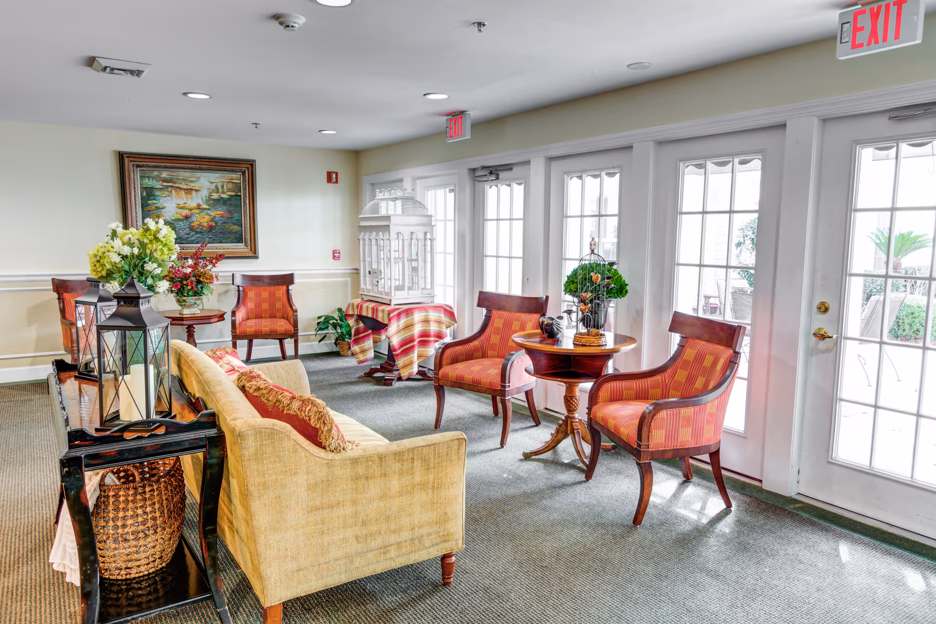 Bright communal lounge with upholstered chairs, a sofa, small tables, decorative plants and large French doors to the outside.
