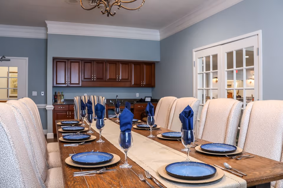 A formal dining room with a long wooden table set for eight people. Each place setting includes a blue plate, a glass with a blue napkin, and silverware. The room has light blue walls, a chandelier, and a set of wooden cabinets along the back wall. There are upholstered chairs around the table and French doors on the right side.