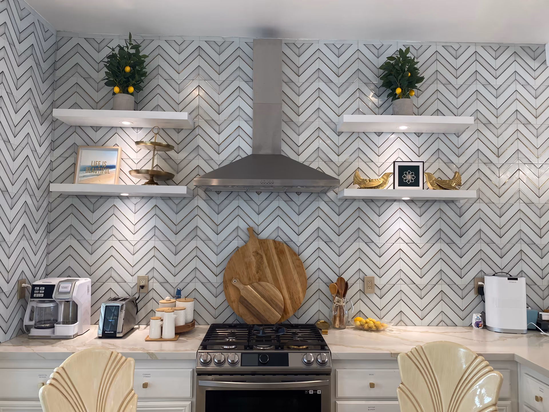 Modern kitchen with chevron tile backsplash, stainless steel range and hood, floating shelves with decor, countertop appliances and bar chairs.