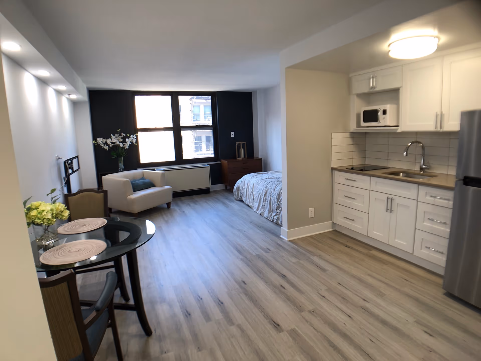 Bright studio apartment interior with a kitchenette on the right, dining table and seating on the left, and a bed by a large window.