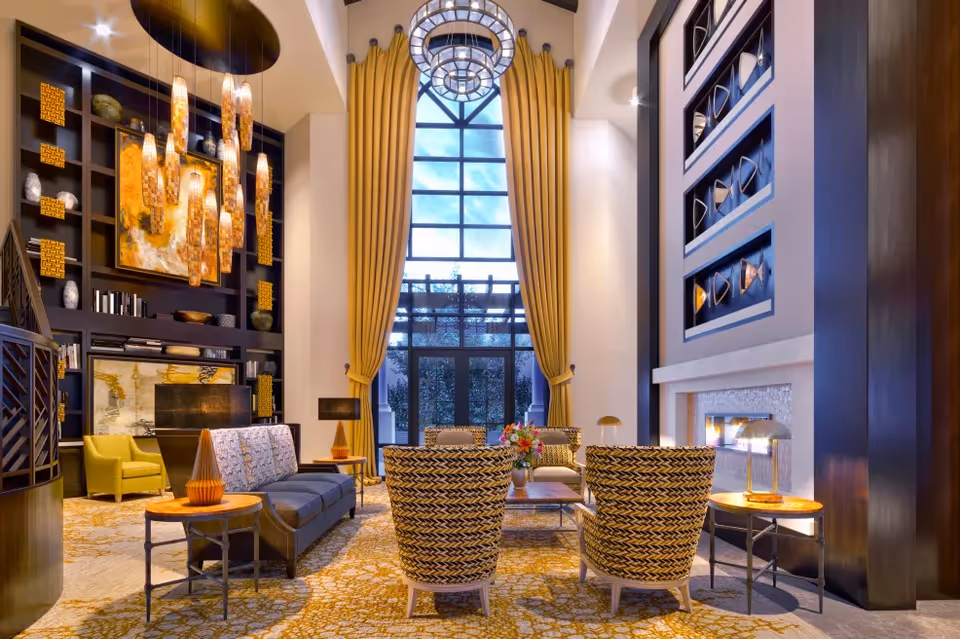Elegant high-ceiling lounge with tall arched window, chandeliers, yellow curtains, seating area, fireplace and decorative shelving.