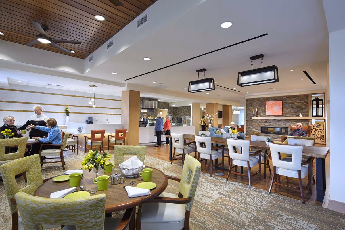A spacious and well-lit common area in a senior living facility featuring a dining section with tables set with green cups and plates, several elderly people sitting and chatting, a modern fireplace with stacked firewood, and a kitchen counter where staff are serving residents.