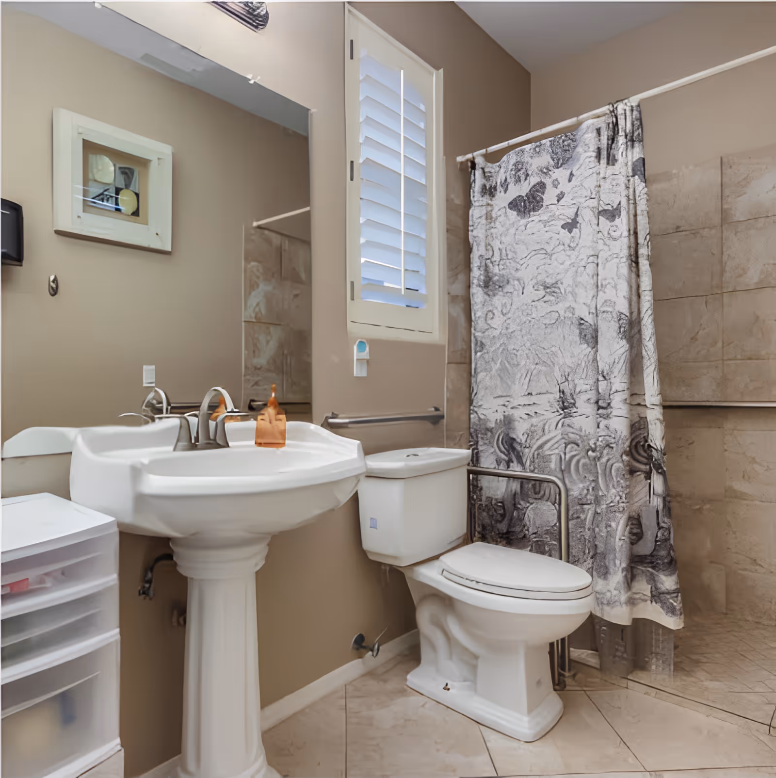 A clean bathroom featuring a white pedestal sink with a faucet, a toilet with a safety grab bar, a shower area with a patterned shower curtain, beige tiled walls and floor, a window with white shutters, and a white plastic storage drawer unit.