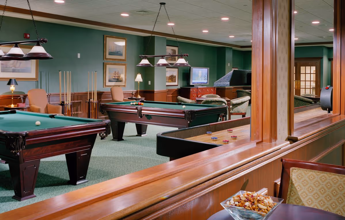 A recreational room with two pool tables, a shuffleboard table, several chairs, and framed paintings on green walls. The room has overhead lights and a television in the background.