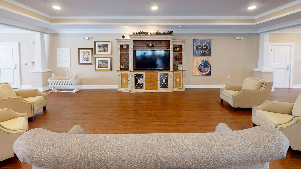 A spacious living room area with wooden flooring, beige walls, and recessed ceiling lights. The room features a large entertainment center with a flat-screen TV, decorative shelves, and framed artwork on the walls. There are several beige upholstered armchairs and a sofa arranged around the room, creating a comfortable seating area.