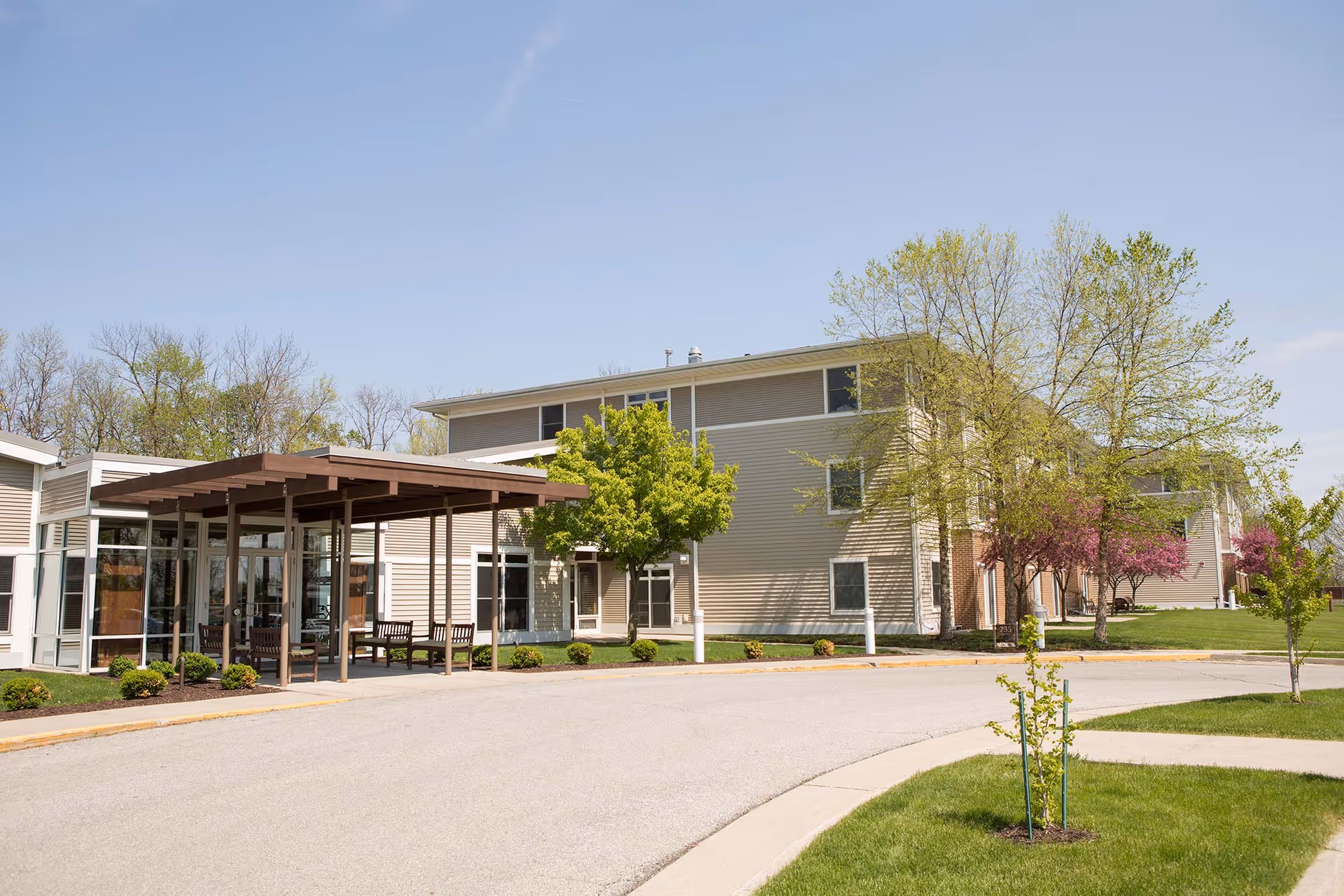 Exterior front entrance of a senior living building with a covered drop-off area, driveway, lawn, and trees.
