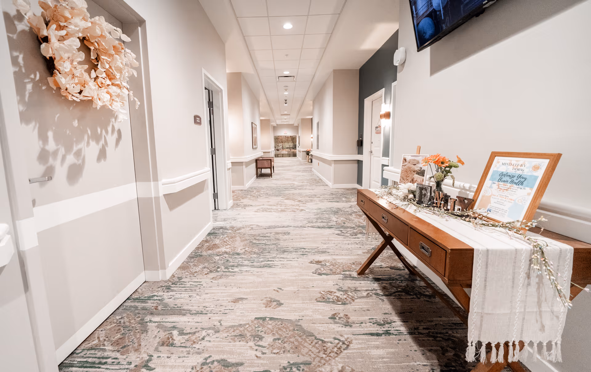 Bright, decorated hallway in an assisted living facility with a wooden console table and doors along the corridor.