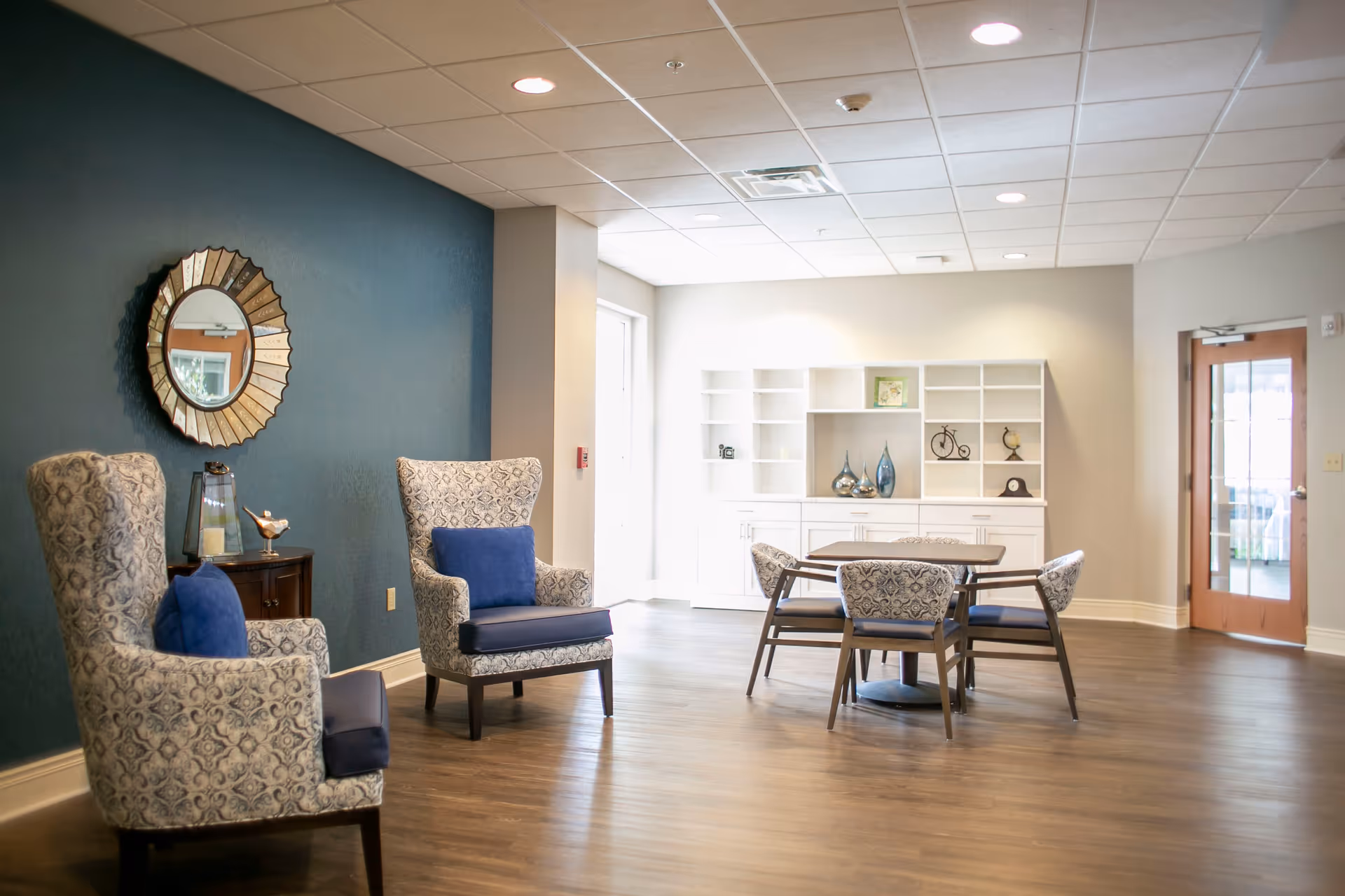 A bright and spacious common area with two patterned armchairs with blue cushions, a small wooden side table with decorative items, a round mirror on a dark teal accent wall, a table with four matching chairs, and a white shelving unit with decorative objects. The room has wooden flooring and a ceiling with recessed lighting.
