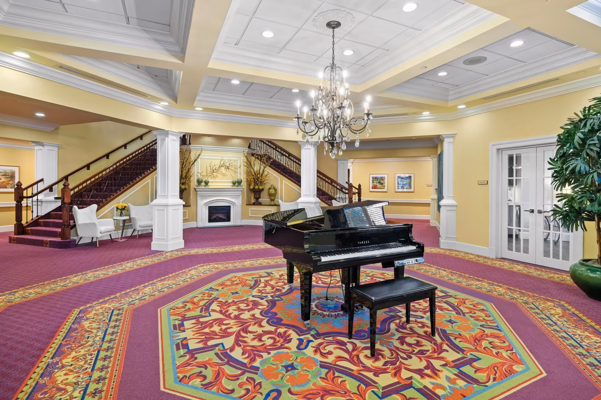 Spacious and elegant lobby area with a grand piano on a colorful patterned carpet, a chandelier hanging from the ceiling, a staircase with wooden railings, white columns, a fireplace, and seating areas with white chairs and small tables.