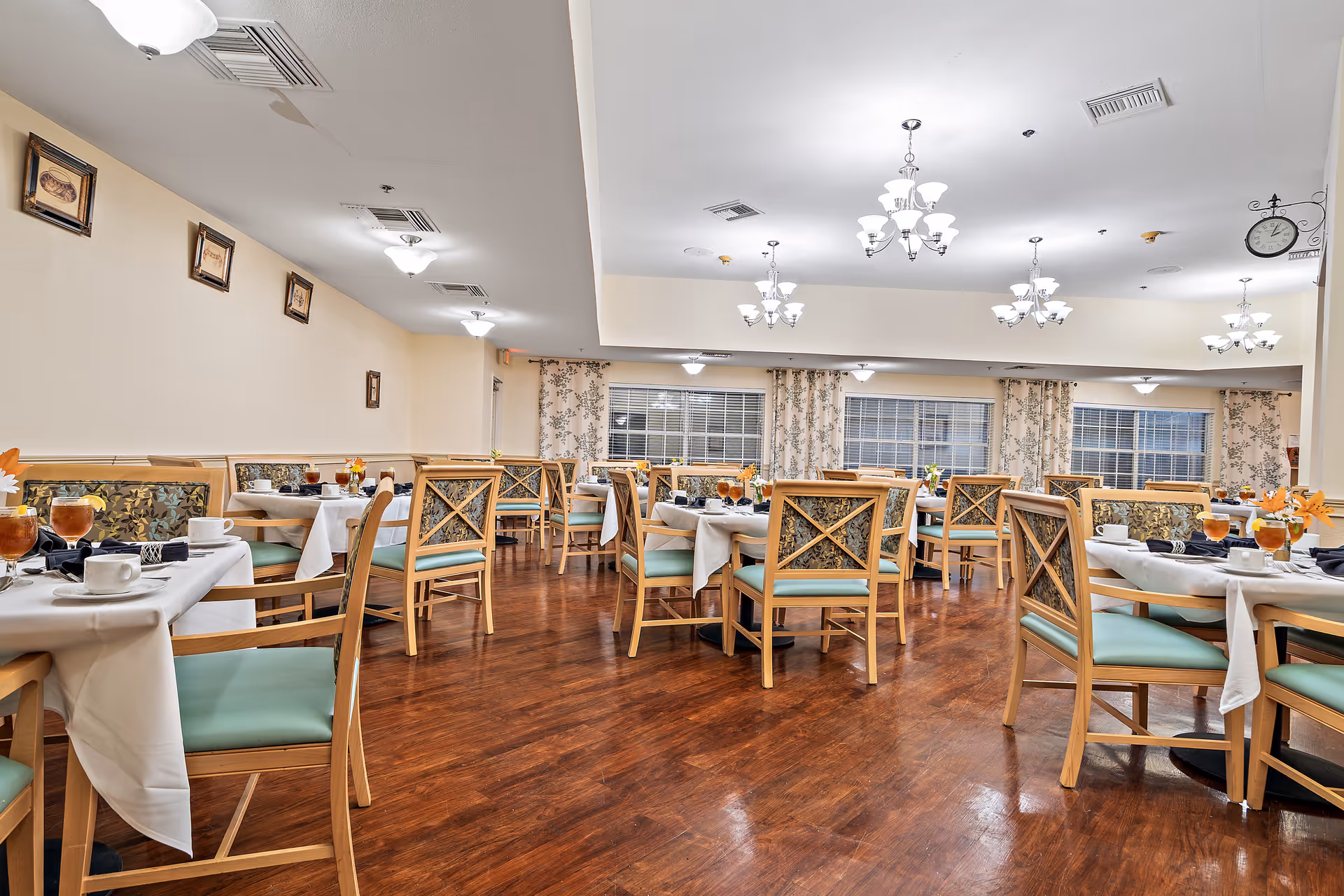 A spacious dining room with multiple tables covered in white tablecloths, set with cups, glasses of iced tea, and floral centerpieces. The room features wooden chairs with light green cushions and patterned backs, hardwood flooring, floral curtains on the windows, and several ceiling light fixtures providing bright illumination.