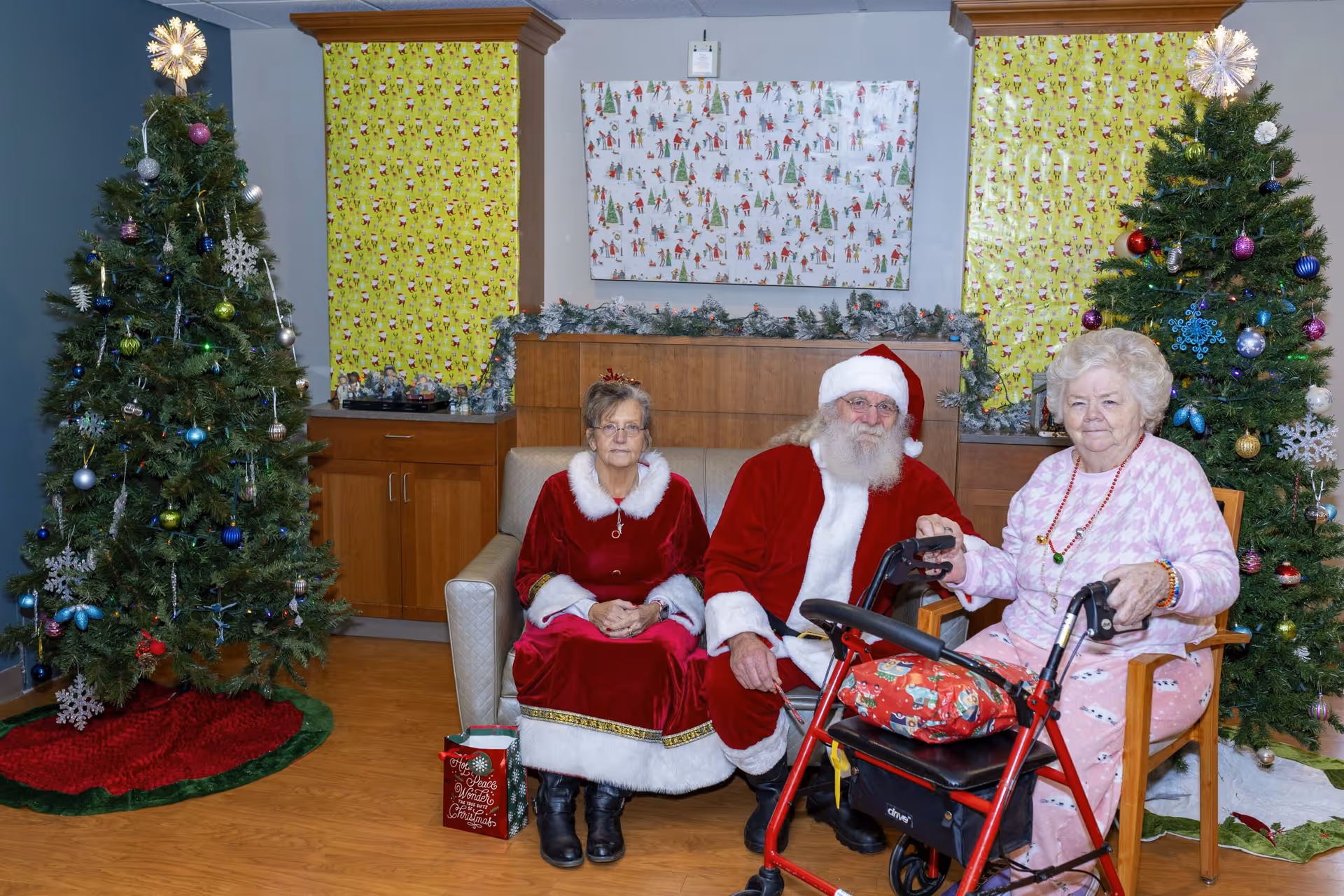Two elderly women and a man dressed as Santa Claus sitting in a room decorated for Christmas with two decorated Christmas trees on either side and festive wrapping paper on the walls.