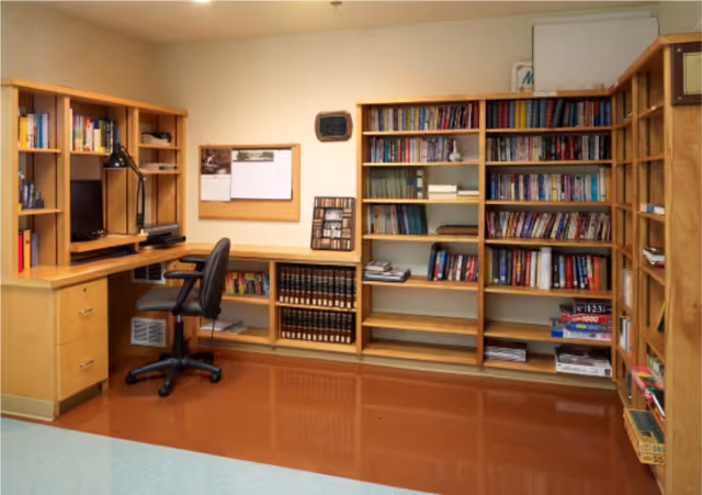 A cozy room with wooden bookshelves filled with books and board games, a wooden desk with a computer monitor and office chair, a cork bulletin board on the wall, and a polished brown floor.