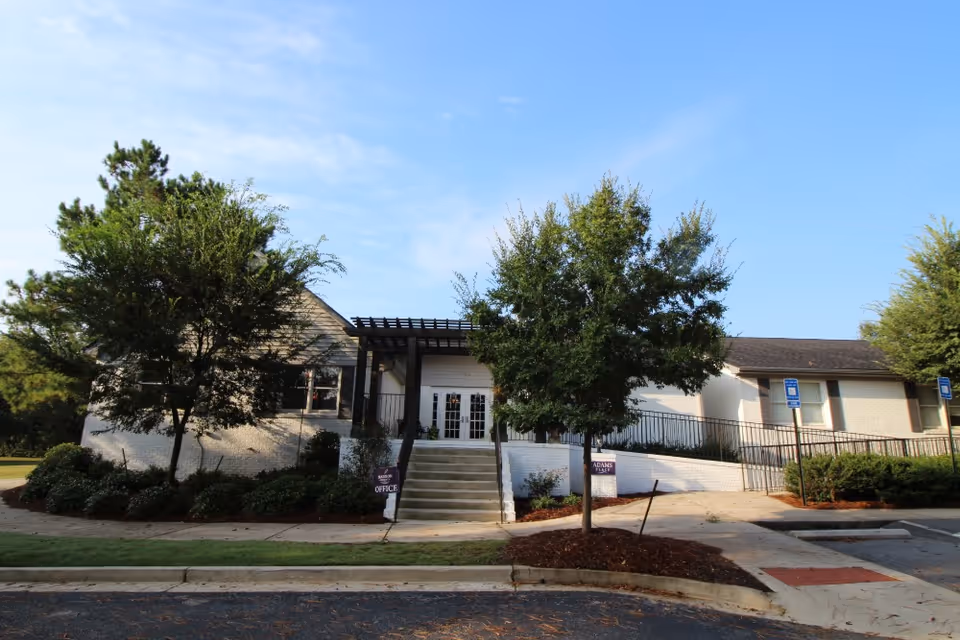 Exterior view of a single-story building with white brick walls and a dark roof, featuring a central entrance with stairs and a ramp on the right side. There are two trees and landscaped bushes in front, along with signs indicating 'Madison Office' and 'Adams Place'. The sky is clear and blue.