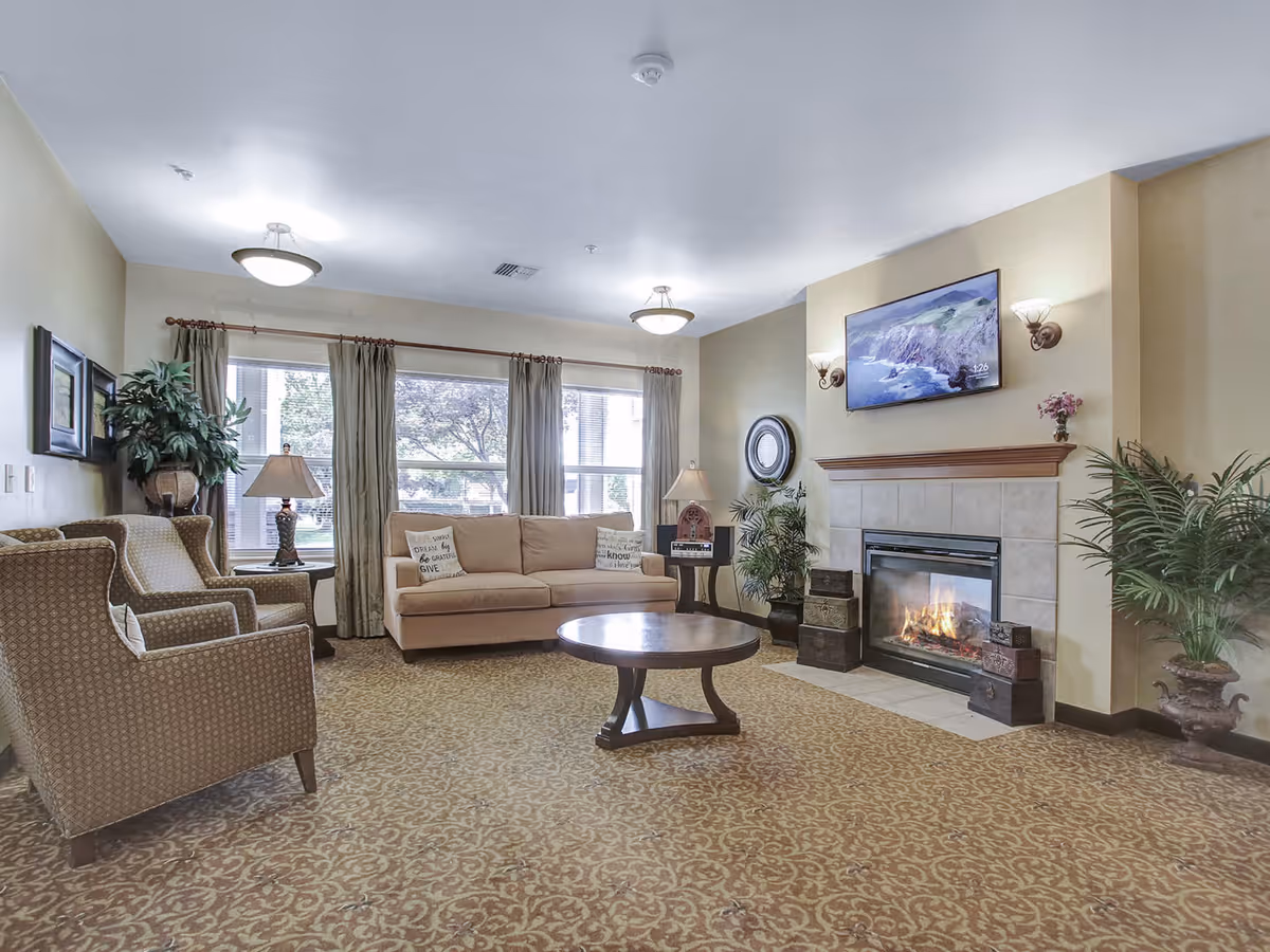 A cozy senior living facility common area with a beige sofa, two patterned armchairs, a round wooden coffee table, and a fireplace with a mounted TV above it. The room has large windows with curtains, decorative plants, wall sconces, and a patterned carpet.