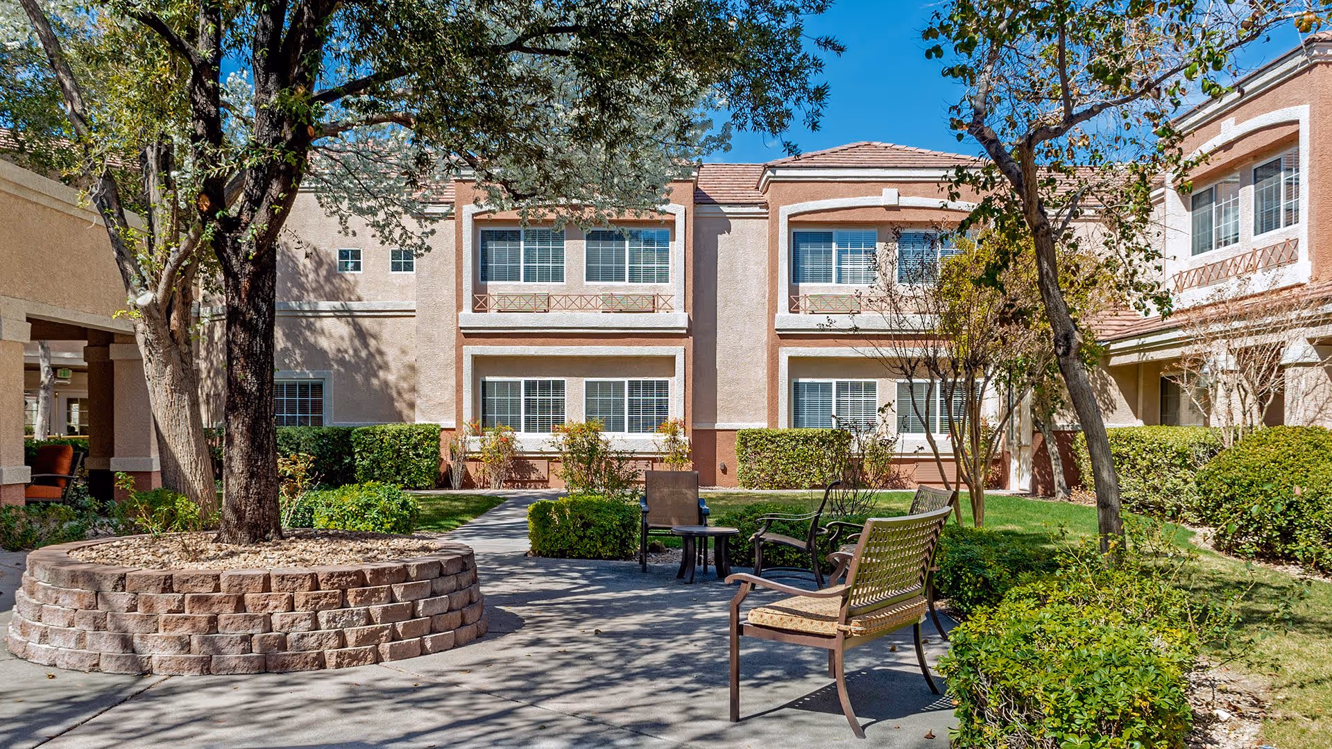 Outdoor courtyard area at Atria Seville with a large tree surrounded by a circular stone planter, several chairs and a small table on a paved area, green bushes, and a two-story building with multiple windows in the background under a clear blue sky.