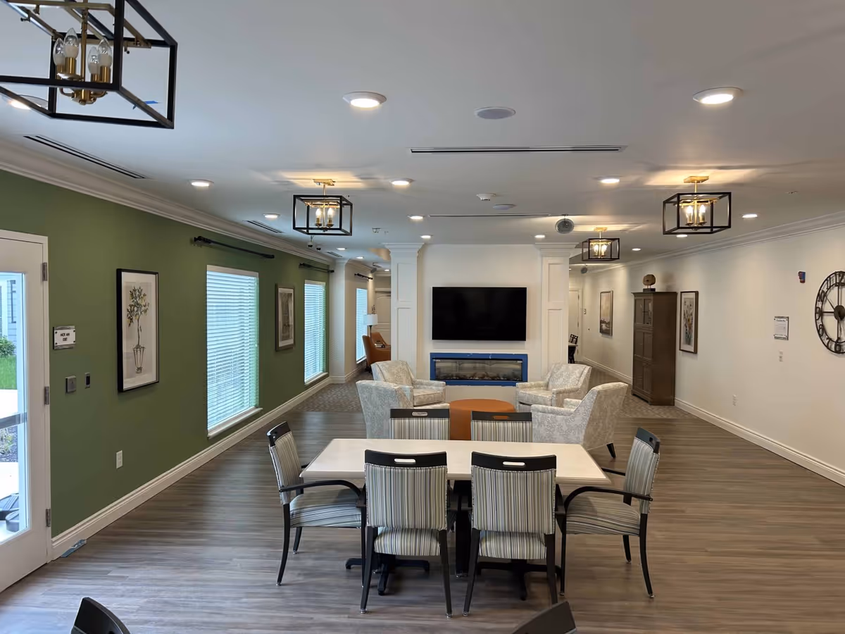Open common living area with a dining table and chairs in the foreground and a seating area with a wall-mounted TV above a fireplace in the background.