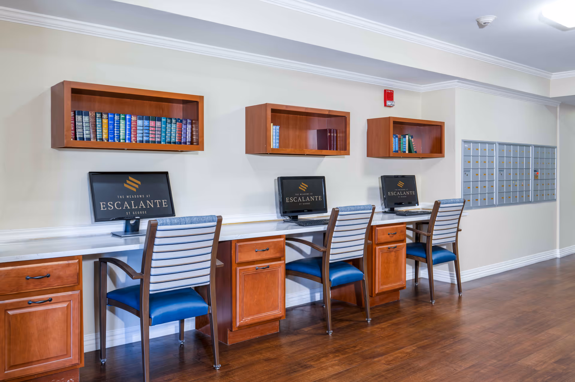 A computer workstation area in a senior living facility with three wooden desks and chairs. Each desk has a computer monitor displaying The Meadows at Escalante logo. Above each desk are wooden wall-mounted shelves with books. To the right, there is a wall of mailboxes. The floor is wooden and the walls are light-colored.