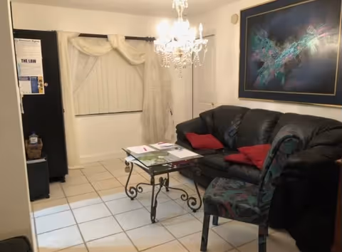 A small living room with a black leather couch adorned with red pillows, a glass coffee table with papers on it, a patterned chair, a chandelier hanging from the ceiling, a large abstract painting on the wall, and a window with vertical blinds and sheer curtains.